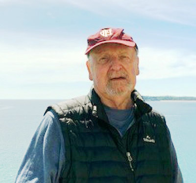 A photograph of Dr Frank Ainsworth. He is wearing a red cap and standing in front of a background of blue sky and blue sea. alt=